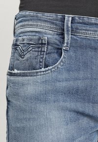 Light blue denim jeans with a faded texture, featuring a front pocket and stitched accents near the waistband. Distressed hem detail visible.