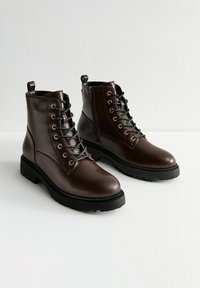 Bottes en cuir marron à la cheville avec un laçage à l'avant, des œillets en métal et une fermeture éclair sur le côté. Semelle en caoutchouc noir épais et finition texturée.