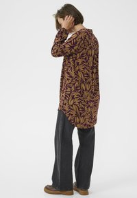 Chemise à manches longues de couleur violet foncé avec un motif de feuilles dorées, présentant un ourlet arrondi. Associée à un jean noir ample et des chaussures marron épaisses.