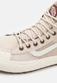 Beige Vans korkean tason tennarit valkoisilla nauhoilla, ruskeilla silmukoilla, pilkullisella valkoisella pohjalla ja valkoisella sivuviivalla nupukkinahkamateriaalissa.