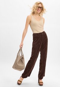 Glunderende vrouw met krullend blond haar, die een bril draagt, een beige mouwloos shirt en een bruine corduroy broek aan heeft, terwijl ze een taupe handtas vasthoudt en bruine platform sandalen draagt.