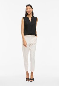 Musta hihaton paita V-pääntiellä, yhdistettynä kermaisiin räätälöityihin housuihin. Malli seisoo mustissa korkokengissä, kädet taskuissa, valkoista taustaa vasten.