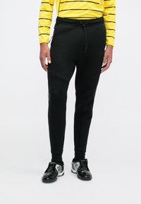 Černé sportovní tepláky s tapered střihem, se šňůrkovým pasem a panelovými detaily. Oblečené s černo-stříbrnými fotbalovými botami.