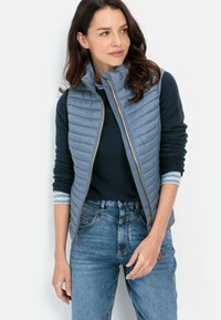 Blau gesteppte Weste mit hohem Kragen, kombiniert mit einem marineblauen Langarmhemd und hellblau gestreiften Manschetten, ergänzt durch blaue Jeans.