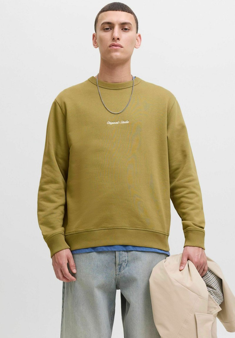 Felpa verde oliva con polsini e orlo a coste, caratterizzata da un logo in bianco. Indossata sopra una camicia blu, mentre tiene in mano un giubbotto beige.