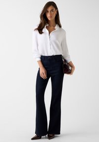 Camicia bianca a bottoni, pantaloni scuri in denim a zampa, scarpe marroni appuntite, che tiene una piccola borsa bordeaux. Presenta tessuto liscio e vestibilità su misura.