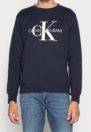 Mężczyzna ubrany w granatową bluzę z białym napisem "Calvin Klein Jeans" i dużym logo "cK", w połączeniu z jasnoniebieskimi jeansami.