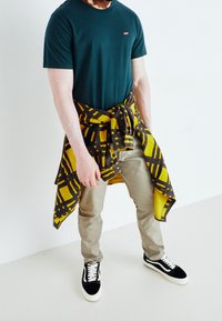 Mand iført mørkegrøn t-shirt, beige bukser, sort-hvide sneakers og en gul-sort ternet skjorte bundet om livet.