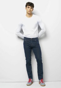 Un jean bleu foncé associé à une chemise blanche à manches longues et des baskets grises avec des lacets rouges, présentant une coupe classique et un design minimaliste.