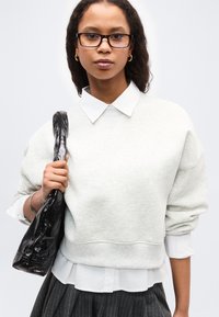 Νεαρή γυναίκα με γυαλιά και χρυσούς κρίκους στα αυτιά κρατά μία μαύρη υφασμάτινη τσάντα, ντυμένη με λευκά ρούχα σε στρώσεις και παντελόνι με ρίγες.