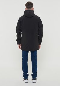 Giacca nera impermeabile con cappuccio, maniche lunghe e silhouette dritta. Indossata con jeans blu e sneaker nere. Design minimalista.