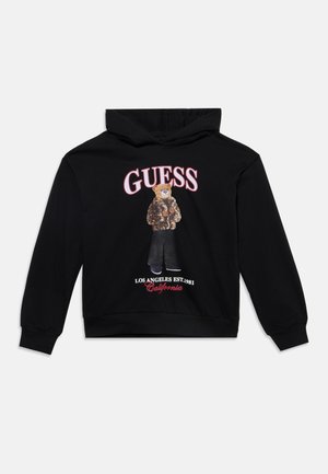 Sweat à capuche noir avec un ours en peluche portant une veste imprimé léopard, un pantalon noir, et le texte "Guess, Los Angeles Est. 1981, California."