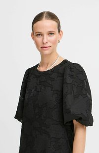 Juoda suknelė su tekstūruota gėlių raštu, trumpomis pūstomis rankovėmis ir apvaliu iškirpimu. Suderinta su sidabriniu grandininiu kaklo papuošalu. Lygus audinys.