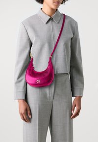 Borsa a spalla in pelle rosa con design curvo, accessori in oro e finitura strutturata, indossata sopra un outfit a scacchi bianco e nero.