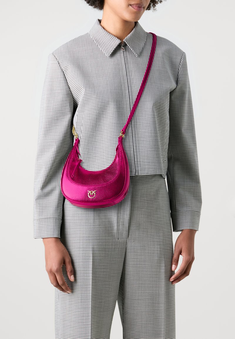 Borsa a spalla in pelle rosa con design curvo, accessori in oro e finitura strutturata, indossata sopra un outfit a scacchi bianco e nero.