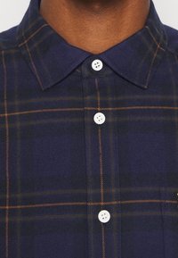 Chemise à carreaux marine en tissu texturé, avec un col boutonné, des boutons blancs et des lignes oranges dans le motif.