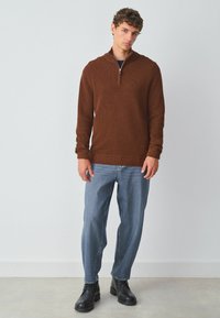 Brauner gestrickter Pullover mit Reißverschluss am Kragen, kombiniert mit hellblauen geraden Jeans und schwarzen Schnürstiefeln. Strukturiertem Stoff und gerippten Bündchen.
