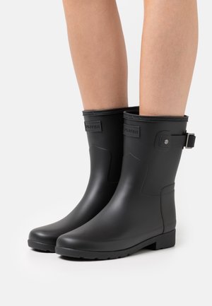 Bottes de pluie en caoutchouc noir avec un talon bas, présentant un bord supérieur texturé, un détail de boucle ajustable et une finition mate.