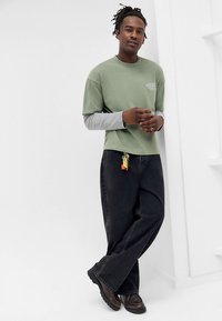 Zielona bawełniana koszulka nałożona na szarą koszulę z długim rękawem, w połączeniu z czarnymi luźnymi dżinsami i brązowymi butami. Brelok przypięty do dżinsów.