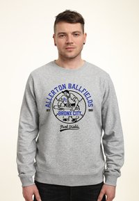 Grijze sweatshirt met ronde hals; voorzien van een bedrukte afbeelding van een honkbalspeler, tekst "Allerton Ballfields Bronx City 1961" in blauw en zwart.