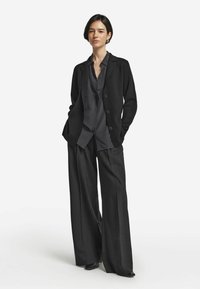 Schwarze maßgeschneiderte Blazer über einem dunkel gestreiften Hemd, kombiniert mit weiten, anthrazitfarbenen Hosen. Das Outfit zeichnet sich durch glatte Texturen und subtile Details aus.