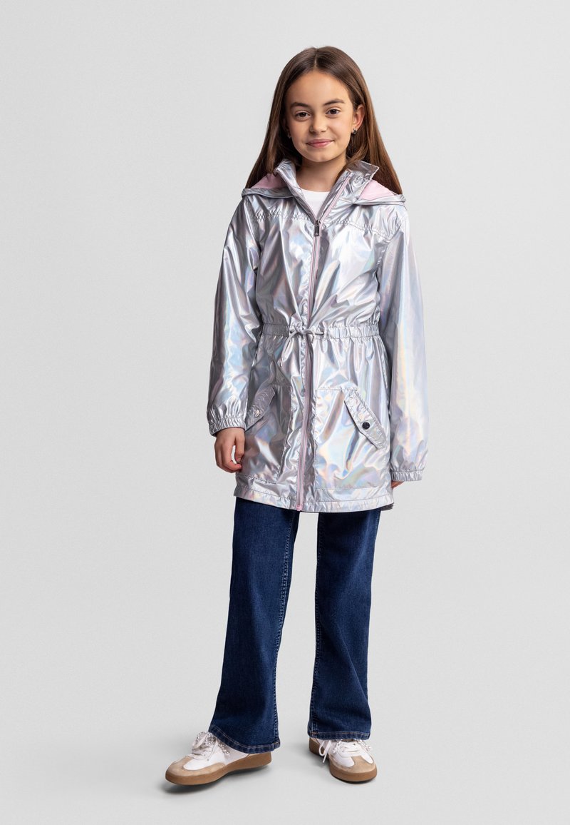 Fille debout portant un imperméable argenté brillant à capuche avec des poches frontales, un jean bleu et des baskets blanches, sur un fond gris uni.