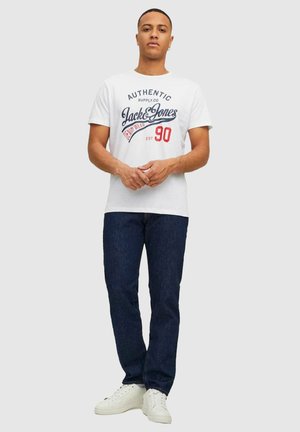 Man wearing white graphic Jack & Jones T-shirt, dark blue jeans, and white sneakers, standing with hands clasped against gray background.