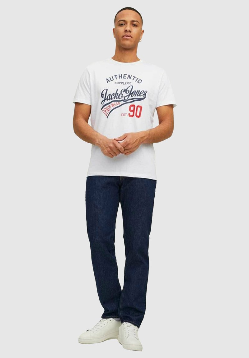 Homme portant un t-shirt blanc graphique Jack & Jones, un jean bleu foncé et des baskets blanches, debout les mains jointes devant un fond gris.