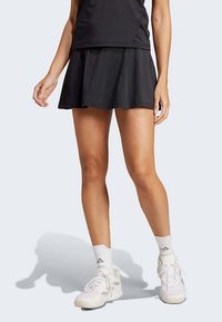 Άτομο που φοράει μια μαύρη αθλητική φούστα, μαύρο τοπ, λευκές κάλτσες Adidas και λευκά παπούτσια Adidas, που στέκεται σε ελαφρύ φόντο.