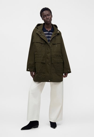 Femme portant une veste à capuche oversize vert olive, un polo rayé bleu marine, un pantalon blanc large et des bottines noires, debout avec une expression neutre.