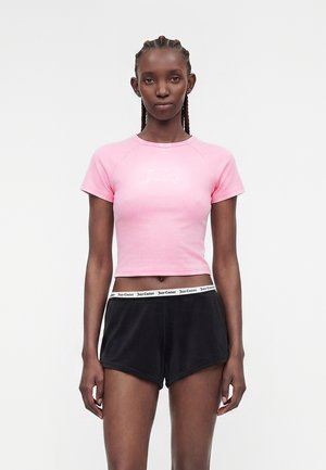 Žena s copánky, která má na sobě růžový přiléhavý krátký top a černé šortky s páskem „Juicy Couture“, stojící na jednobarevném světlém pozadí.