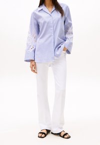 Camicia a maniche lunghe a righe blu e bianche con dettagli ricamati sulle maniche, abbinata a pantaloni larghi bianchi e sandali neri con cinghie.
