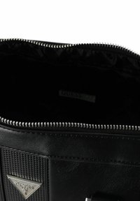 Zwarte synthetische handtas met ritssluiting; heeft gestructureerde strepen, metalen beslag en een zichtbaar logolabel aan de binnenkant. Binnenkant is gevoerd met zwarte stof.