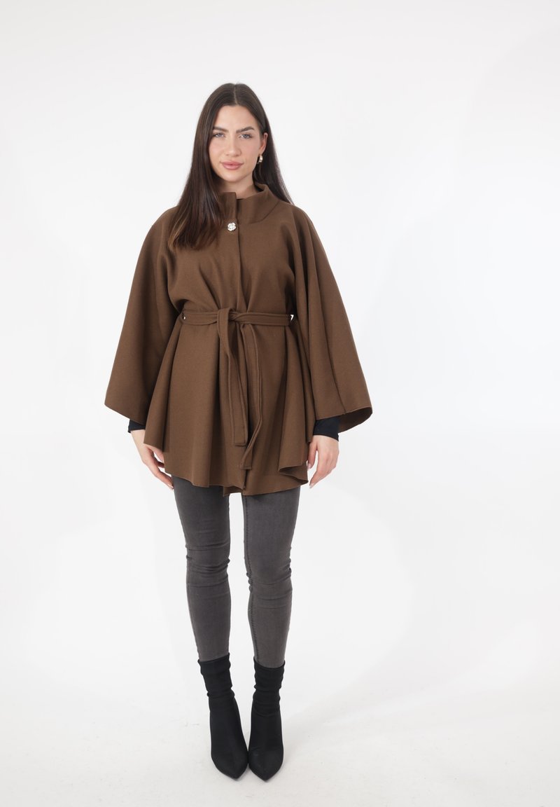 Cappotto marrone con cintura, collo alto e maniche larghe, indossato sopra una maglia nera e jeans skinny grigi, abbinato a stivaletti neri.