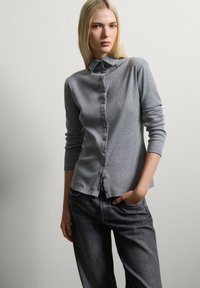 Grauer Knopf-Cardigan mit langen Ärmeln, aus strukturiertem Gewebe. Verfügt über einen Kragen und Knöpfe auf der Vorderseite, kombiniert mit dunklen Jeans.