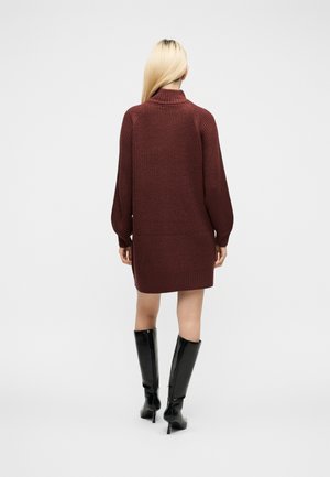 Femme aux longs cheveux blonds portant une robe-pull en tricot bordeaux et des bottes noires montantes à talons, debout, de dos, sur fond blanc.