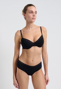 Woman wearing a black bra and matching black underwear, standing against a plain light gray background, looking to her right.