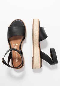 Svarta lädersandaler med öppen tå, plattform och ankelremmar, med vävda beige mellansulor, visade från ovan och sidan på en vit bakgrund.