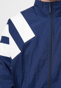 Könnyű anyagból készült tengerészkék dzseki fehér, szögletes csíkokkal a vállon és magas gallérral, elülső cipzáras záródással.