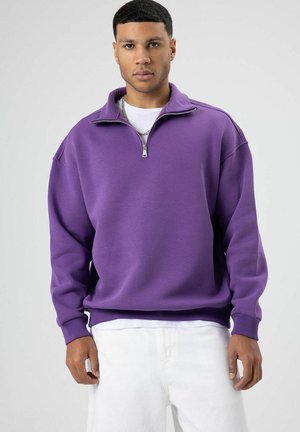 Sweat-shirt en polaire violet avec col zippé quart, coupe décontractée et poignets côtelés. Associé à un pantalon en denim blanc, mettant en valeur un style contrasté.