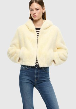 Donna che indossa una giacca soffice color crema sopra una maglia a righe bianco e nero e jeans blu su uno sfondo chiaro e uniforme.