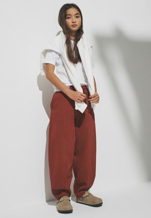Young woman with long hair wears white T-shirt, white sweatshirt draped over shoulders, rust-colored wide pants, and beige slip-on shoes.