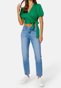 Top verde corto avvolgente con maniche a sbuffo legato in vita, abbinato a jeans a vita alta azzurri chiari e sneakers bianche.