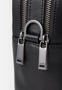 Sac en cuir noir texturé doté d'une double fermeture éclair en métal avec des tirettes rectangulaires, un logo en relief et un accent en cuir lisse.