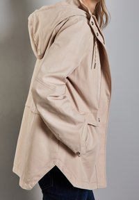 Beige huvjacka med avslappnad passform, som har tryckknappsstängning, sidofickor och en något böjd fåll. Slät, matt textur.