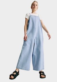 Lichtblauwe denim jumpsuit met wijde pijpen, verstelbare bandjes en een rechte snit. Draagt over een wit shirt, gecombineerd met zwarte sandalen.