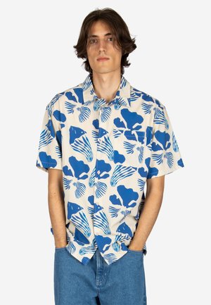 Junger Mann mit mittelbraunem Haar, der ein kurzärmliges beiges Hemd mit blauen abstrakten Mustern und blaue Denim-Jeans trägt.