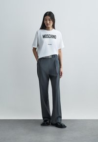 Bílý bavlněný tričko s potiskem "Moschino Milano", kombinované se šedými kalhotami s vysokým pasem a černými botami. Hladká textura a uvolněný střih.