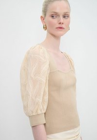 Top beige lavorato a maglia con maniche puff trasparenti ricamate con motivi a foglia, collo alto e silhouette aderente.