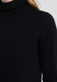 Pull à col roulé noir en tissu doux, avec une coupe ajustée et une texture côtelée au niveau du col et des poignets.